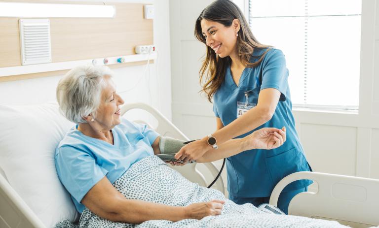 Nurse talking to senior woman in hospital bed. She is measuring her blood pressure.