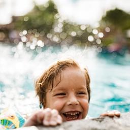 A happy child wearing water wings holding the edge of an outdoor pool on a sunny day.