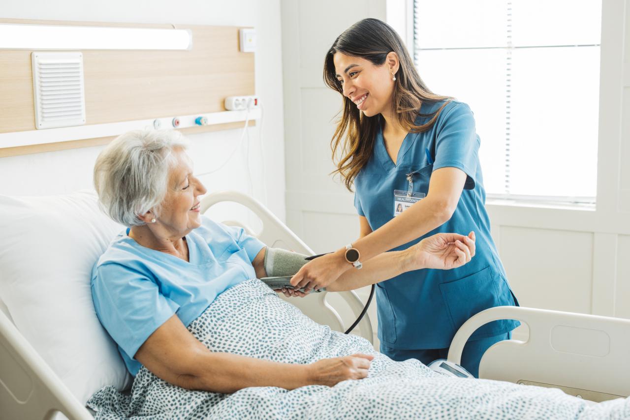 Nurse talking to senior woman in hospital bed. She is measuring her blood pressure.