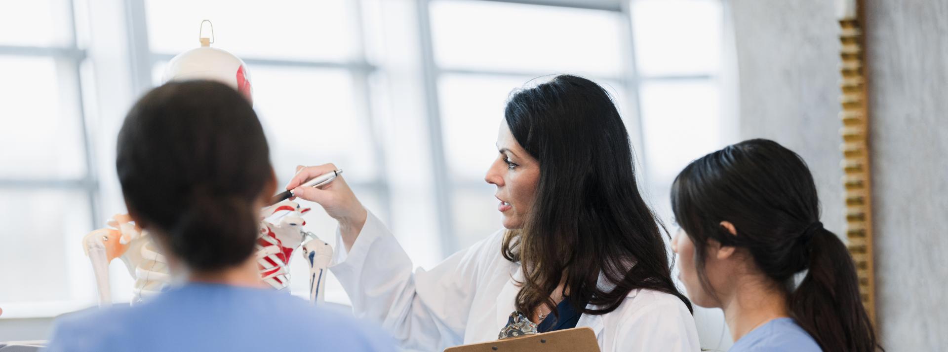 A teacher in a white lab coat points to a model skeleton while two students watch.