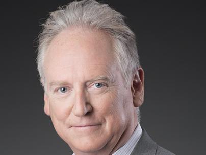 A portrait of John Hedley McLaughlin in front of a grey background.  He is wearing a white patterned shirt and grey blazer
