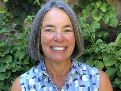 A portrait of Kathy Greenberg in front of green plants.  She is wearing a blue and white patterned top