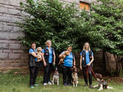 The canines for care team posing with their dogs in front of a brick wall and trees